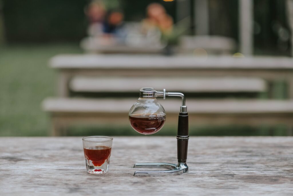 Close-up of a siphon coffee maker and coffee cup on a wooden table outdoors.